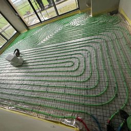 USŁUGI HYDRAULICZNE Filip Turo - Zielone rury ogrzewania podłogowego ułożone na folii z siatką w jasnym pomieszczeniu z dużymi oknami. Widoczne elementy montażowe i fragment nogi osoby.