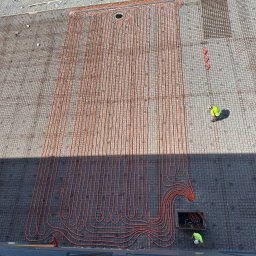 USŁUGI HYDRAULICZNE Filip Turo - Montaż ogrzewania podłogowego w Malborku. Widok z góry na rozłożone rury grzewcze na siatce zbrojeniowej. Pracownik w kasku przy studzience.