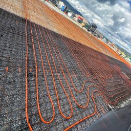 USŁUGI HYDRAULICZNE Filip Turo - Pomarańczowe rury ogrzewania podłogowego ułożone na siatce zbrojeniowej na szarym podłożu żwirowym. W tle budowa, góry i pochmurne niebo.