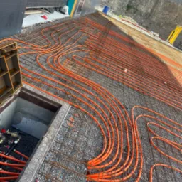 Pomarańczowe rury ogrzewania podłogowego ułożone na siatce zbrojeniowej, widoczne elementy przyłączeniowe w studzience, tło stanowi skalista ściana.