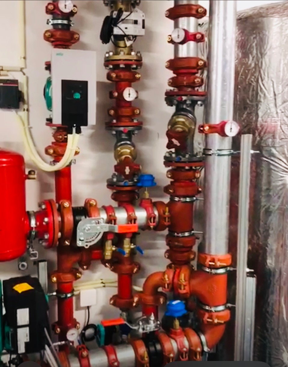 Złożony system rur i zaworów w kolorze czerwonym i srebrnym na białej ścianie, z elementami izolacji. Widoczne manometry i urządzenia sterujące.