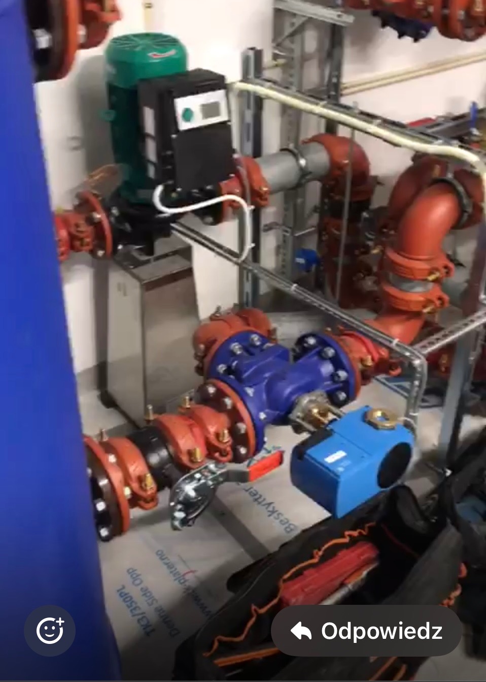 Złożona instalacja hydrauliczna z rurami w kolorze czerwonym i niebieskim, z widocznym silnikiem z zieloną obudową i elementami sterującymi. W tle metalowa konstrukcja wsporcza.