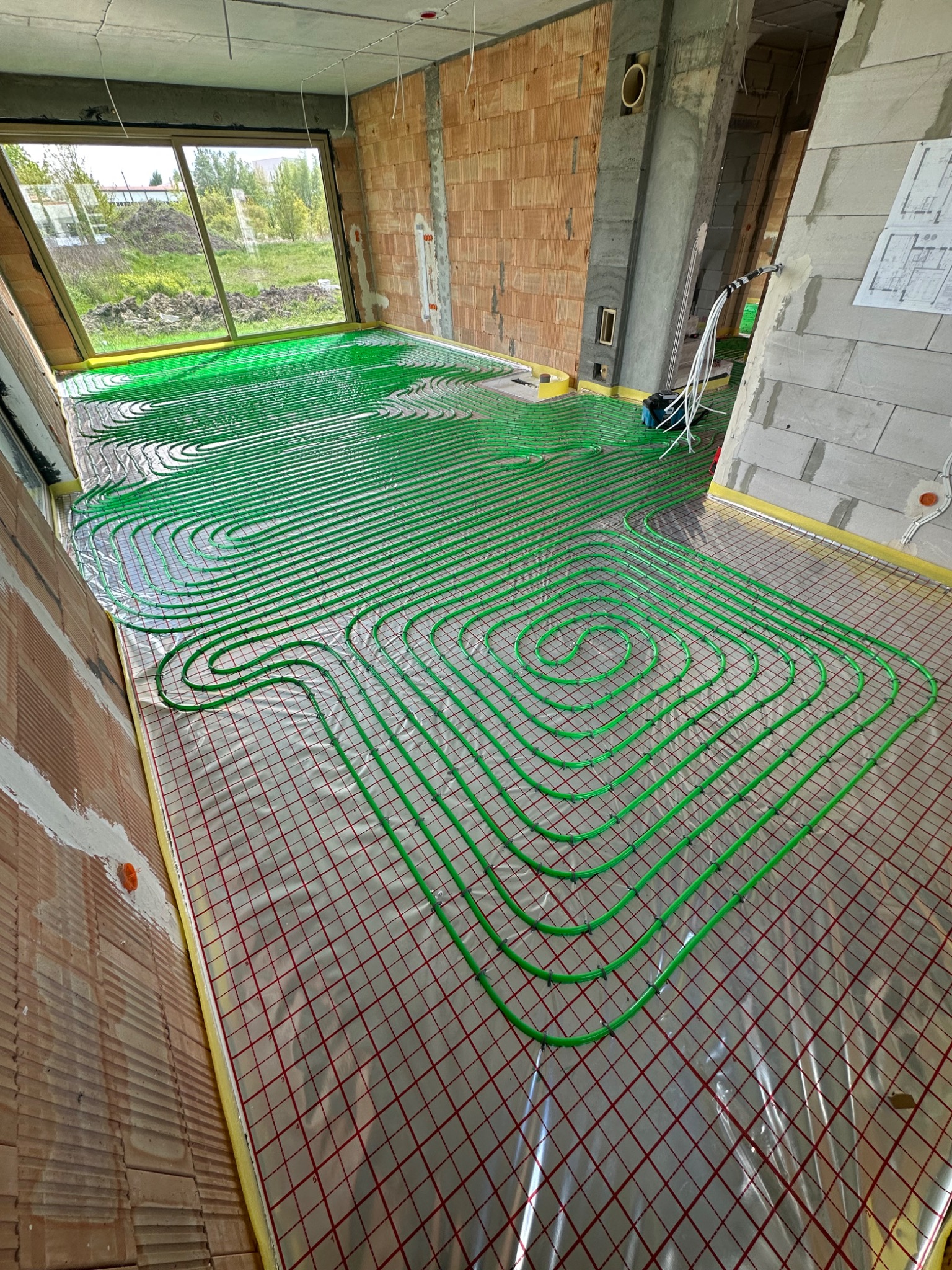 Zielone rury ogrzewania podłogowego ułożone na folii z rastrem w surowym wnętrzu. Widoczne ściany z cegły i okno z widokiem na zieleń. Etap instalacji przed wylewką.