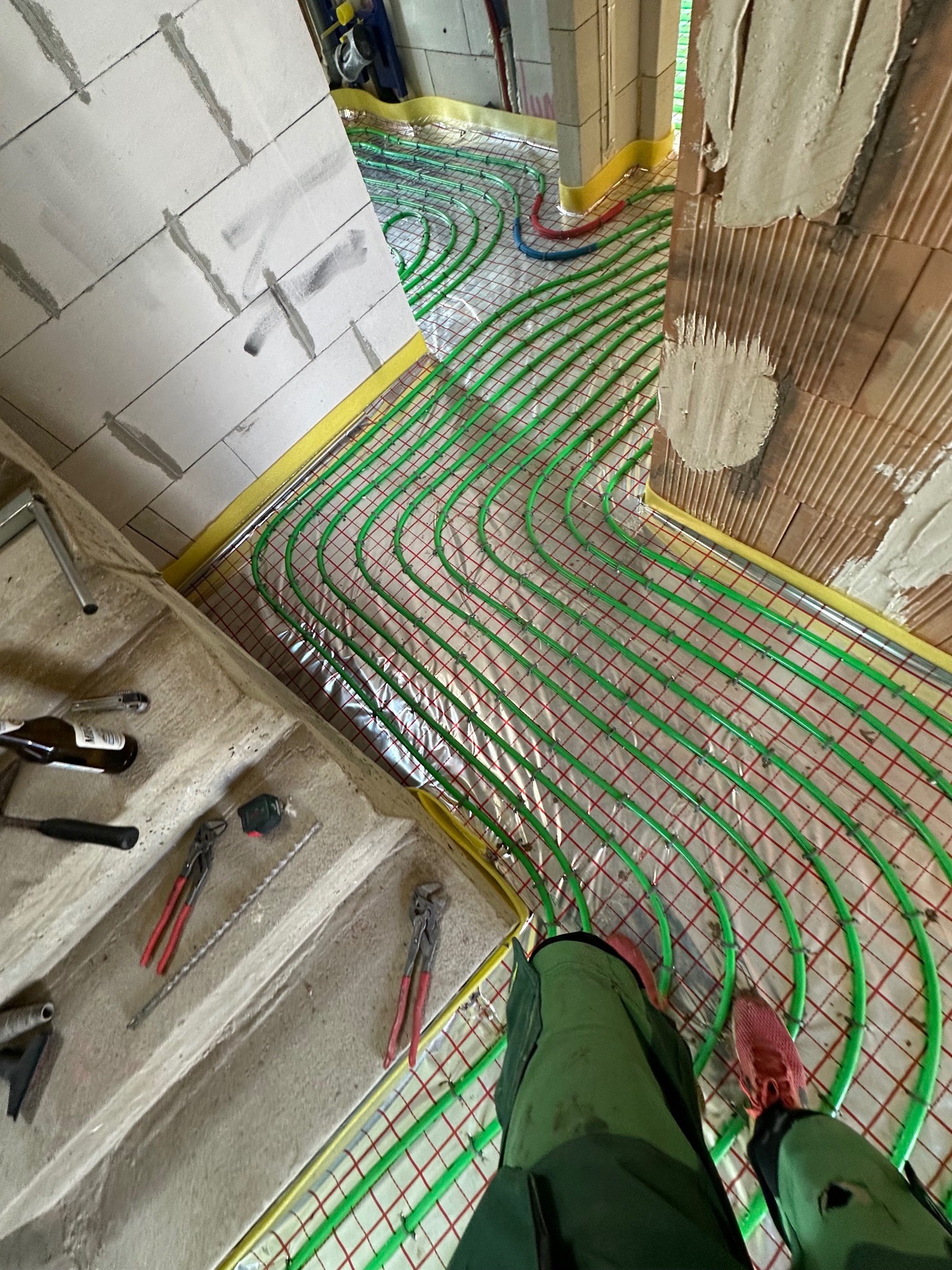 Zielone rury ogrzewania podłogowego ułożone na folii, widoczne narzędzia hydrauliczne i fragmenty ścian w surowym stanie. Żółta taśma na obrzeżach.