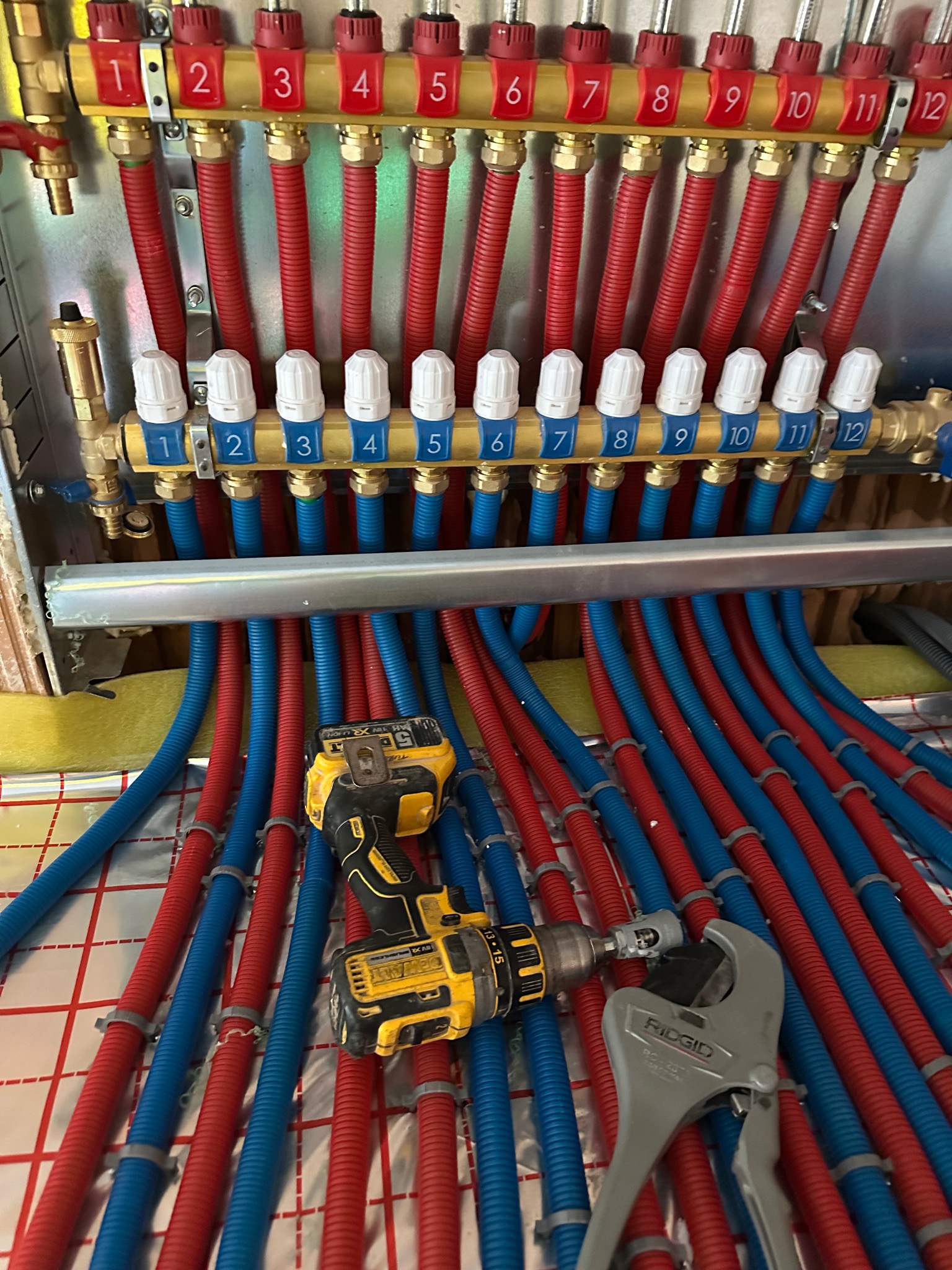 Rozdzielacz ogrzewania podłogowego z rurami PEX (czerwone, niebieskie) i narzędziami (wiertarka, klucz) na folii izolacyjnej, Malbork.