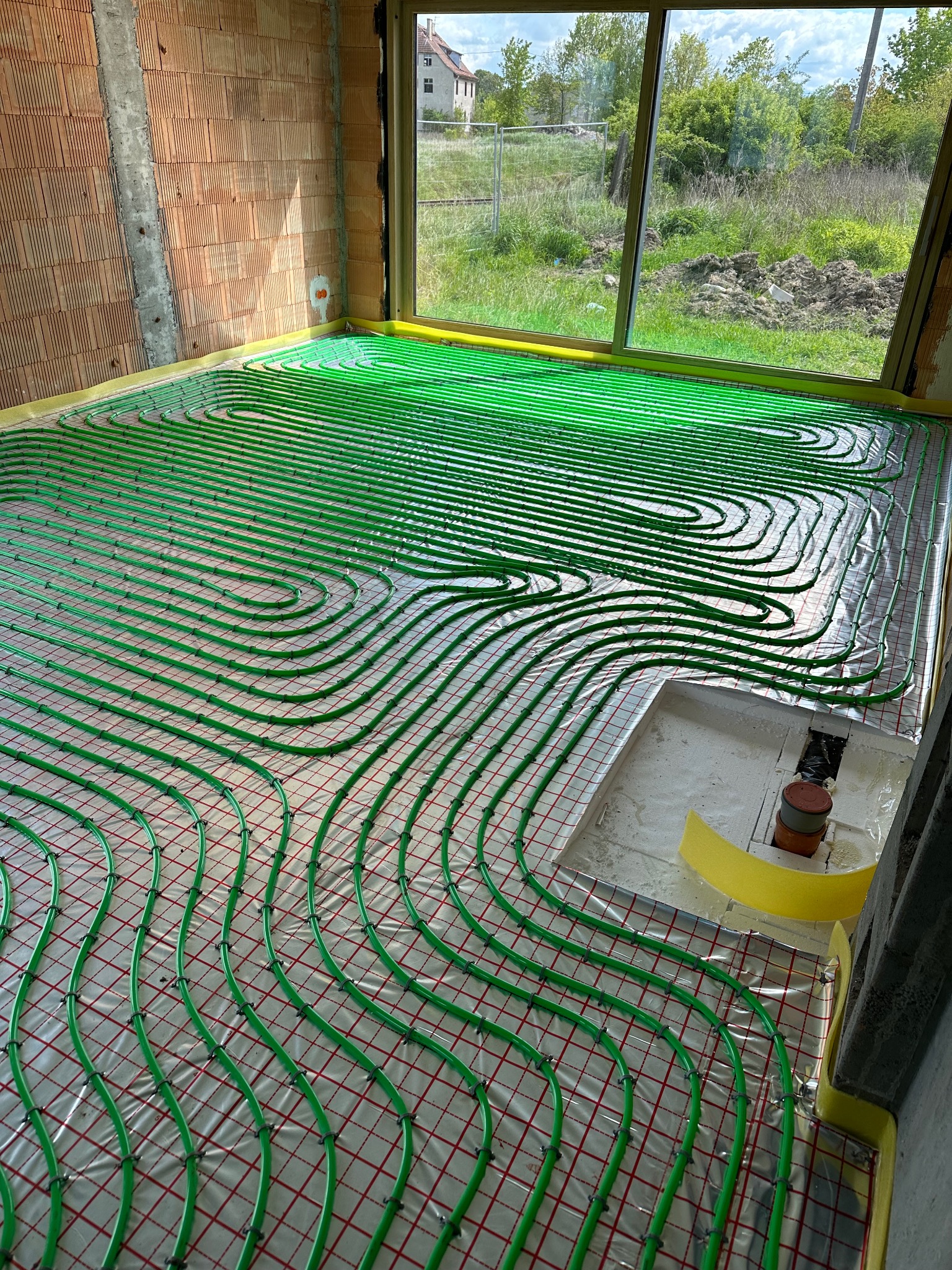 Zielone rury ogrzewania podłogowego ułożone na folii w surowym wnętrzu z oknem na zewnątrz. Widoczne mocowania rur i fragmenty instalacji. Ściany z cegły.