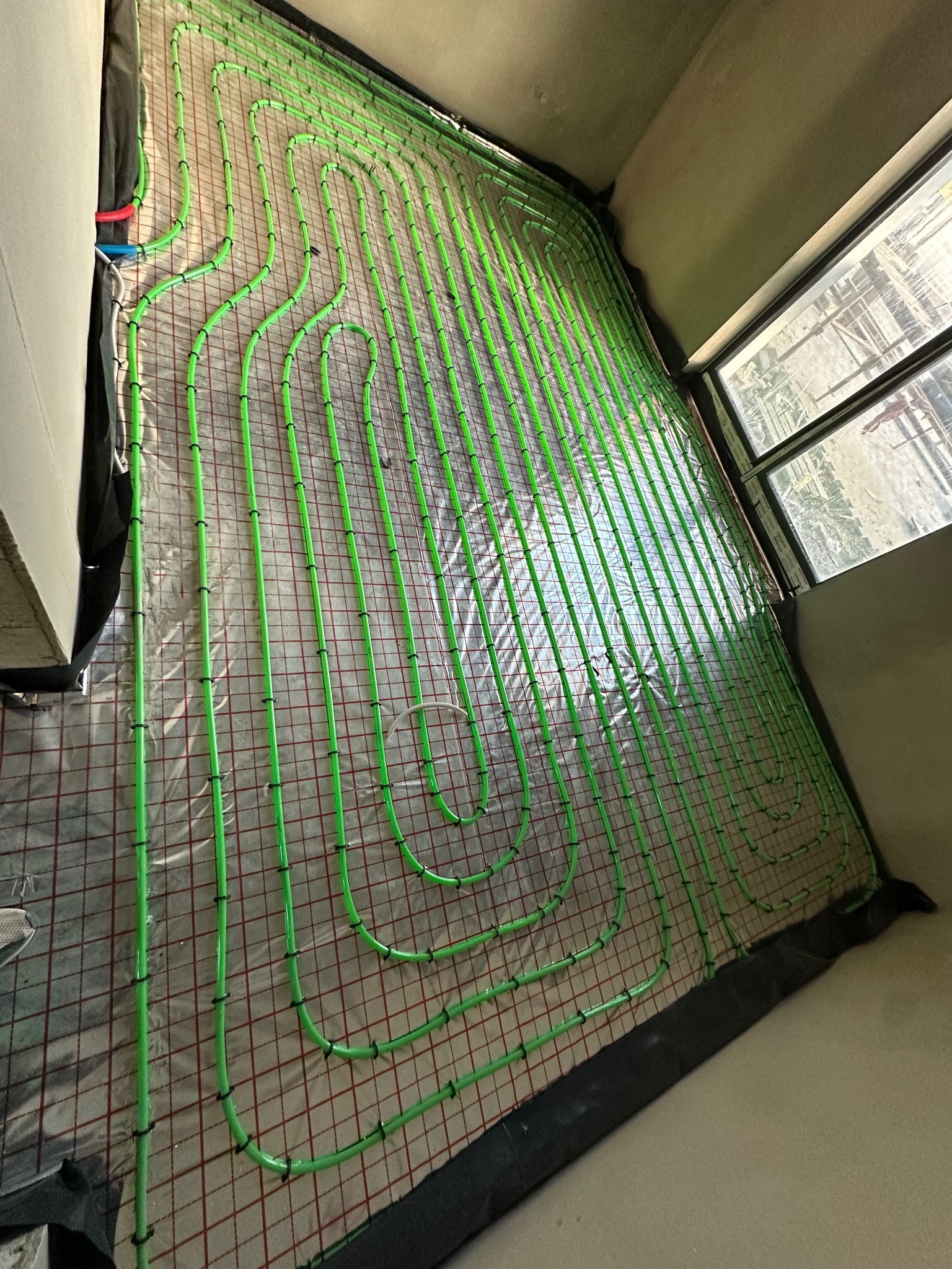 Zielone rury ogrzewania podłogowego ułożone na folii z czerwoną siatką w surowym wnętrzu z oknem. Widok z góry, rury przymocowane czarnymi uchwytami.