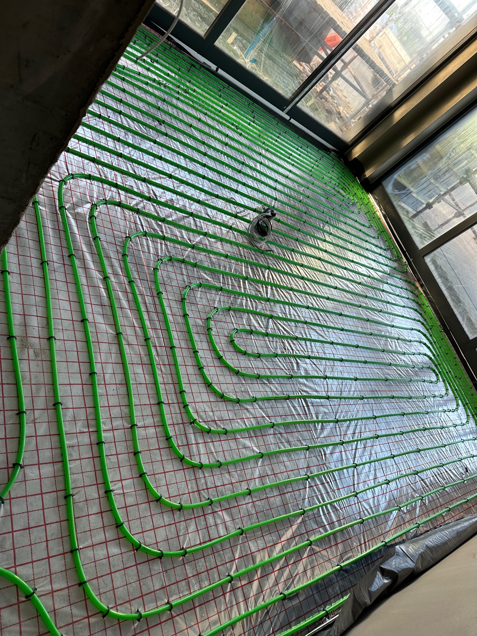 Zielone rury ogrzewania podłogowego ułożone na folii z czerwoną siatką, widoczne okna i fragment budowy w tle, rury przymocowane czarnymi uchwytami.