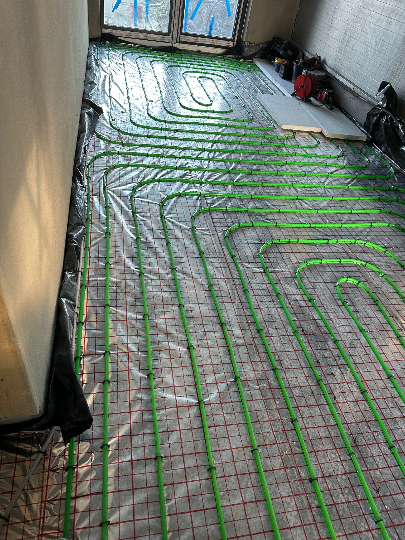 Zielone rury ogrzewania podłogowego ułożone na folii z czerwoną siatką w surowym wnętrzu. Widoczne okno i narzędzia w tle. Instalacja przed wylewką.