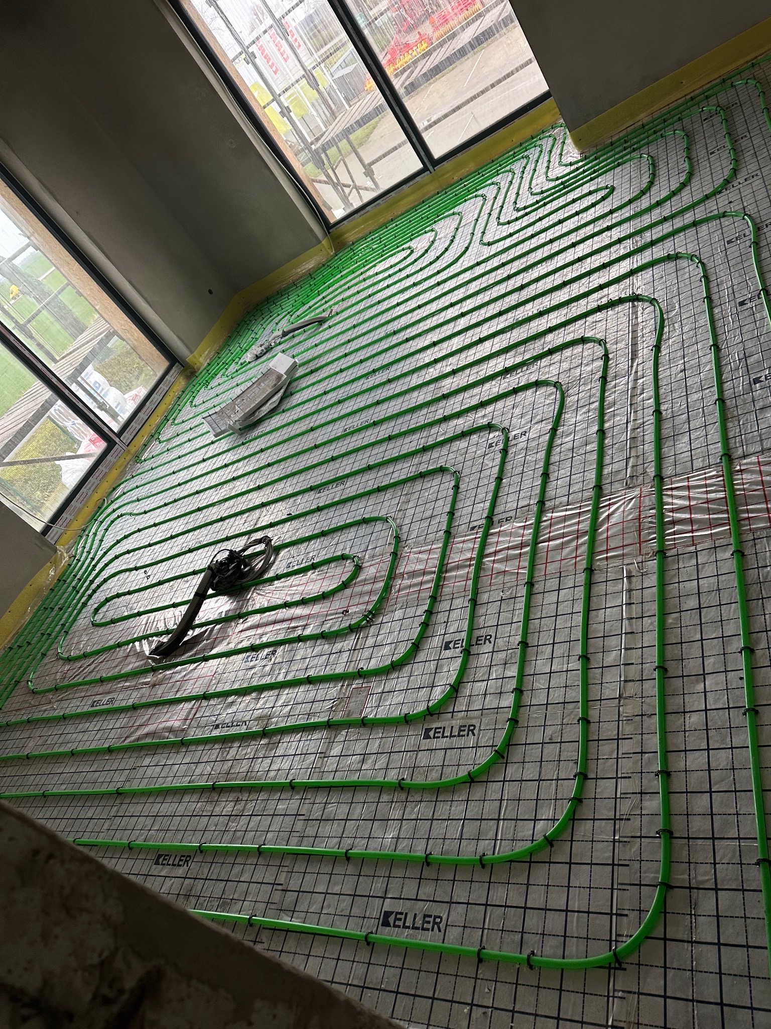 Zielone rury ogrzewania podłogowego ułożone na izolacji z siatką, widoczne okna i narzędzia. Instalacja w trakcie montażu, przed zalaniem posadzki.