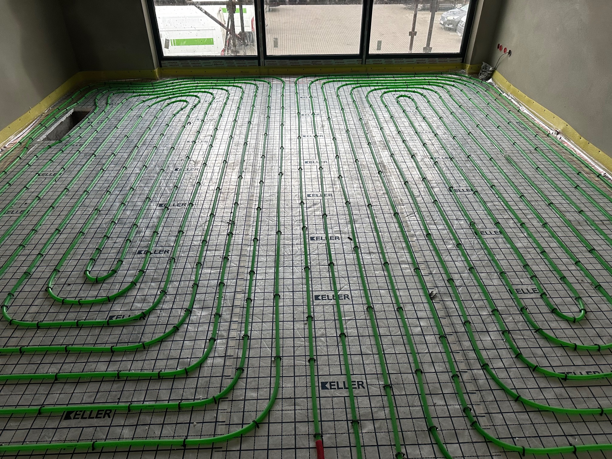 Zielone rury ogrzewania podłogowego ułożone na czarnej siatce, widok z góry, przygotowane do zalania posadzką, żółta taśma brzegowa przy ścianach.