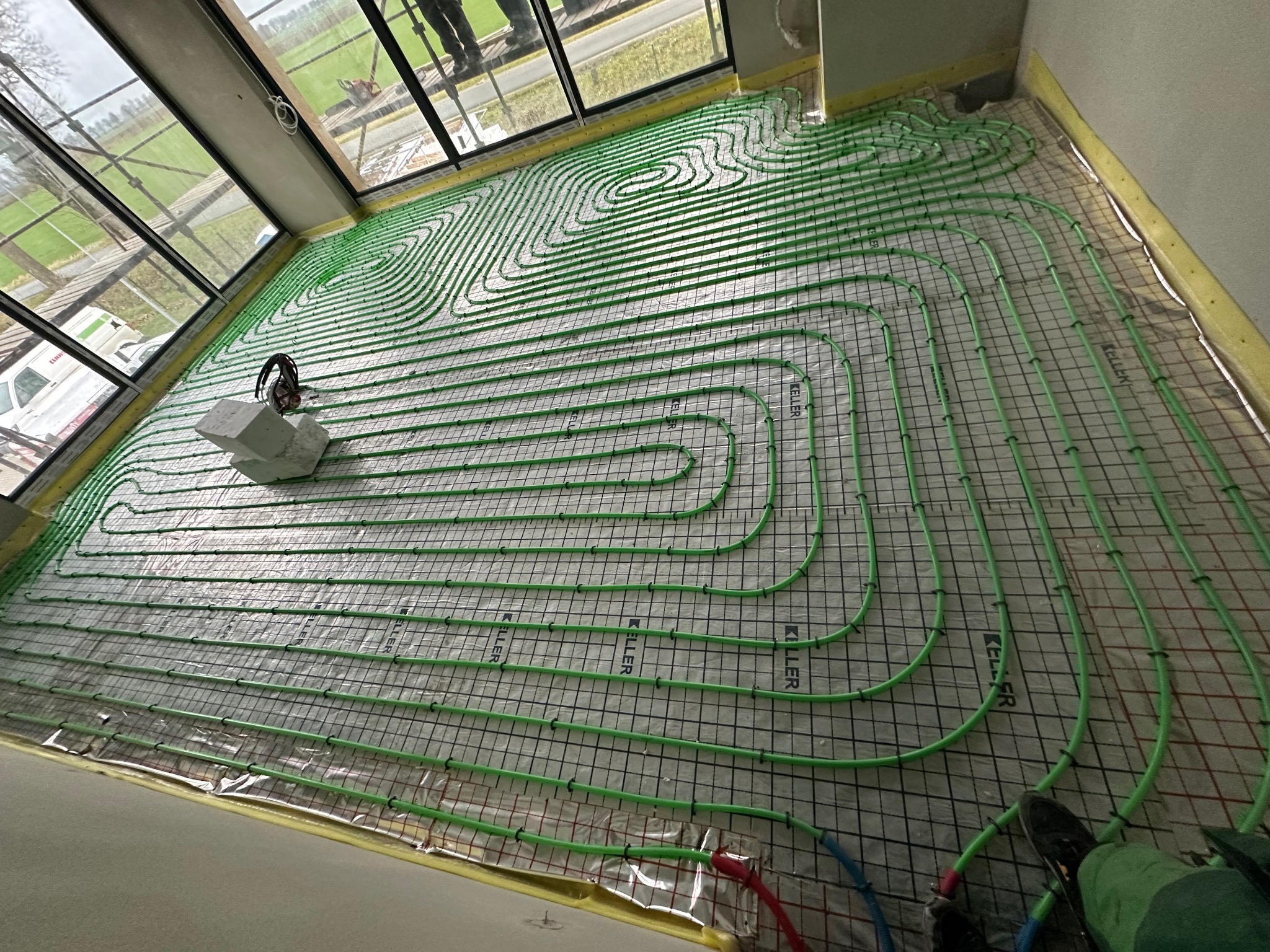 Zielone rury ogrzewania podłogowego ułożone na folii z siatką w jasnym pomieszczeniu z dużymi oknami. Widoczne elementy montażowe i fragment nogi osoby.