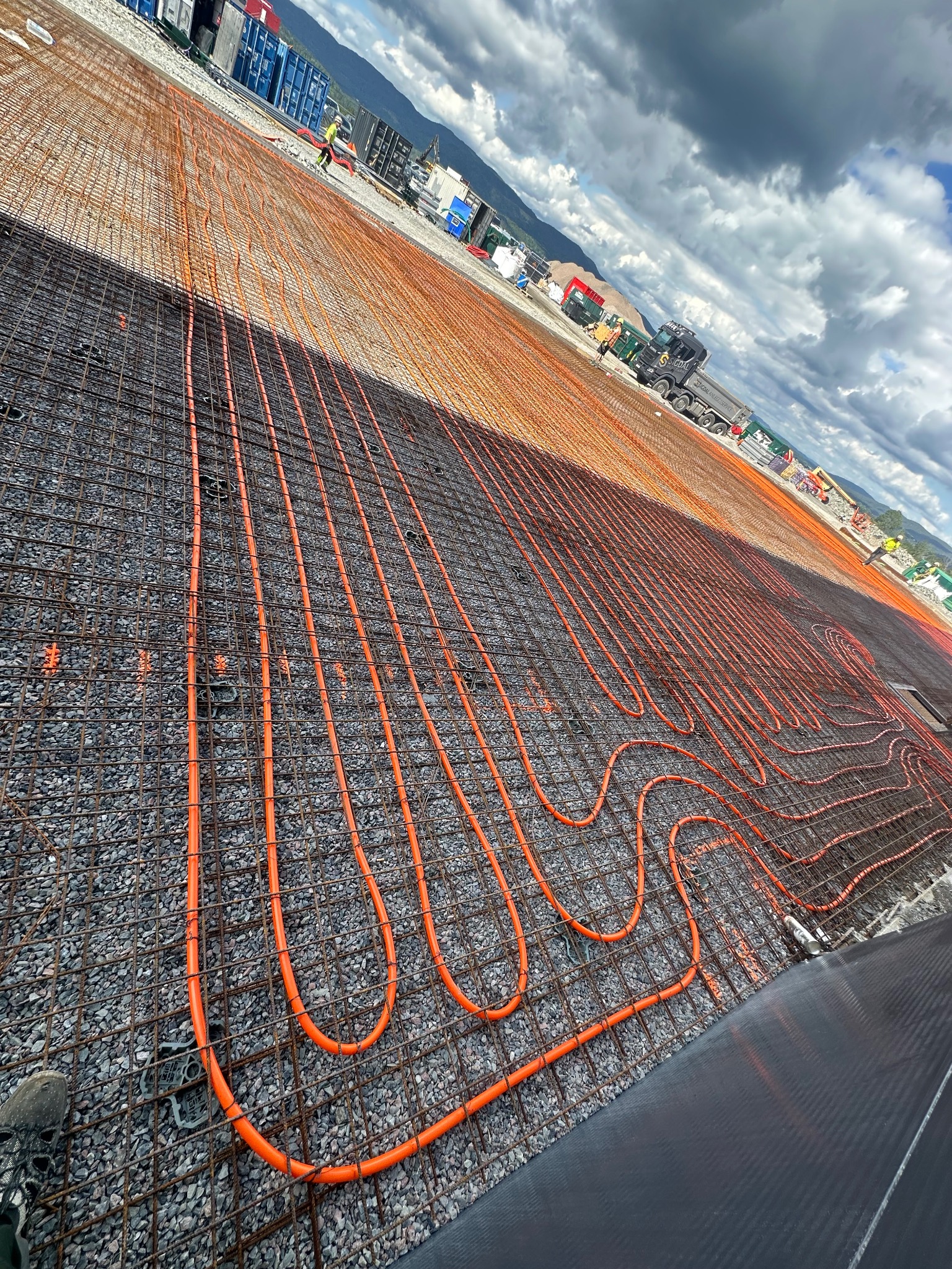 Pomarańczowe rury ogrzewania podłogowego ułożone na siatce zbrojeniowej na szarym podłożu żwirowym. W tle budowa, góry i pochmurne niebo.