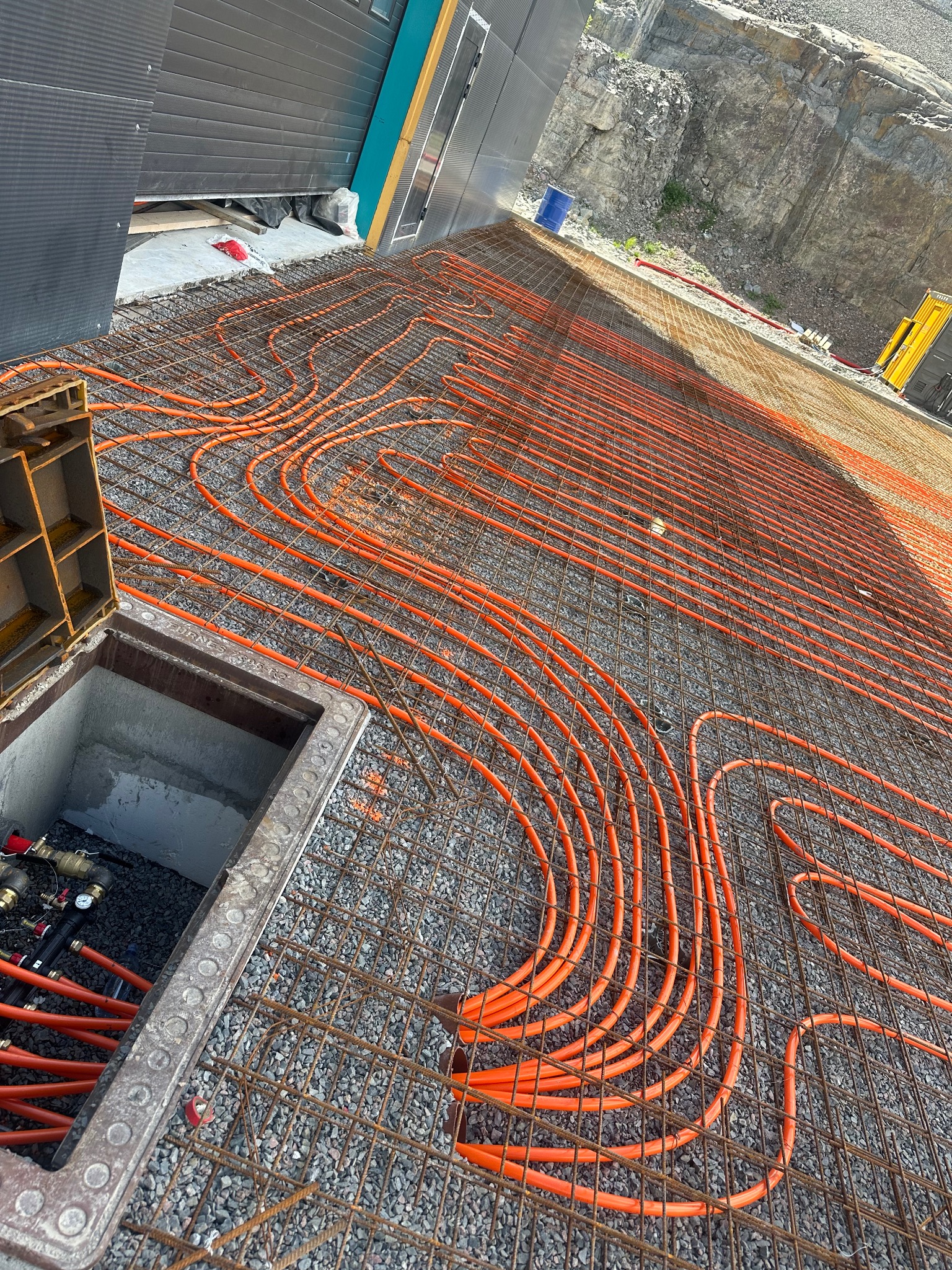 Pomarańczowe rury ogrzewania podłogowego ułożone na siatce zbrojeniowej, widoczne elementy przyłączeniowe w studzience, tło stanowi skalista ściana.