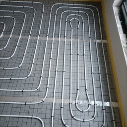 STN Uladzimir Nikitashyn - Białe rury ogrzewania podłogowego ułożone na szarej macie z czarną siatką, zabezpieczone taśmą, widok z góry, fragment okna po prawej.