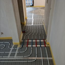 STN Uladzimir Nikitashyn - Rozłożone rury ogrzewania podłogowego na izolacji z siatką w korytarzu. Rury z czerwonymi i niebieskimi oznaczeniami przy ścianach zabezpieczonych taśmą. Widok z góry na instalację.