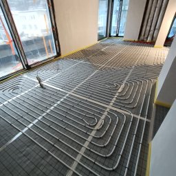 STN Uladzimir Nikitashyn - Białe rury ogrzewania podłogowego ułożone na szarej macie w jasnym pomieszczeniu z dużymi oknami, przygotowane do zalania posadzką.