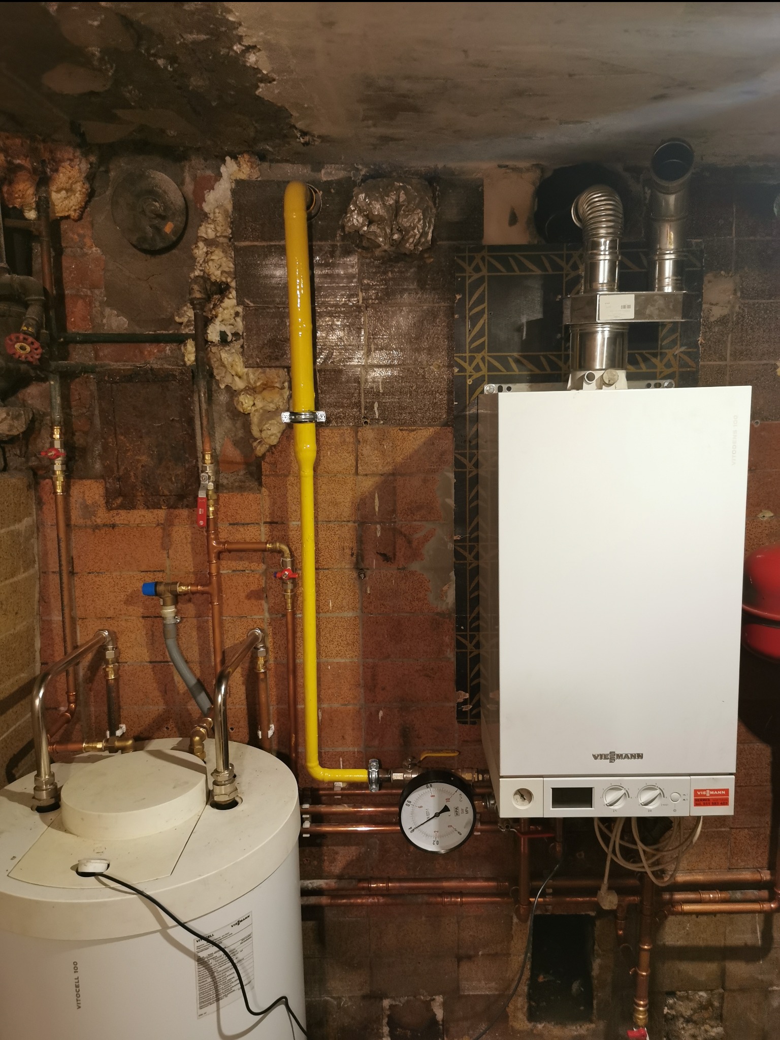 Biały piec gazowy Viessmann z żółtą rurą gazową i bojlerem Vitocell 100 na tle ceglanej ściany z widoczną instalacją hydrauliczną.