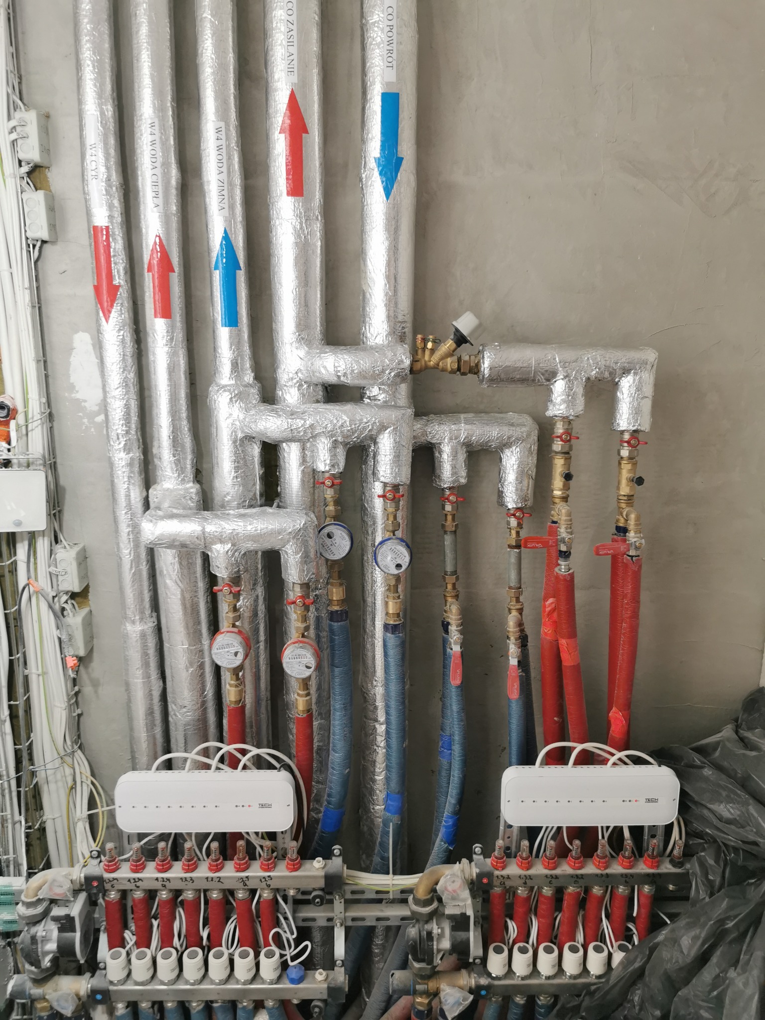 Rozdzielacz ogrzewania podłogowego z rurami oznaczonymi strzałkami (czerwona - zasilanie, niebieska - powrót) na tle surowej ściany. Widoczne liczniki i zawory.