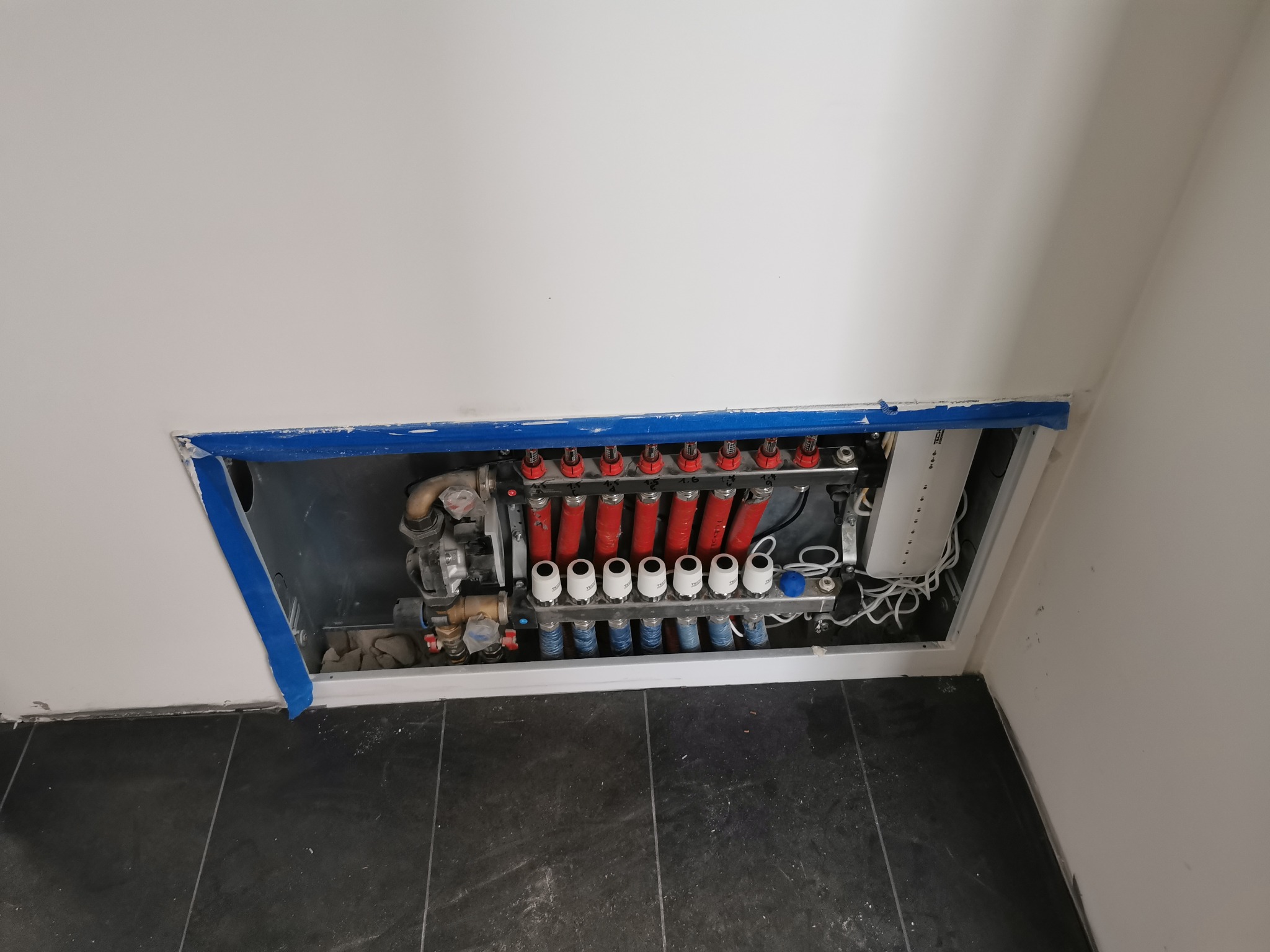 Rozdzielacz ogrzewania podłogowego z czerwonymi rurami i białymi termostatami, zamontowany w ścianie, zabezpieczony niebieską taśmą malarską.