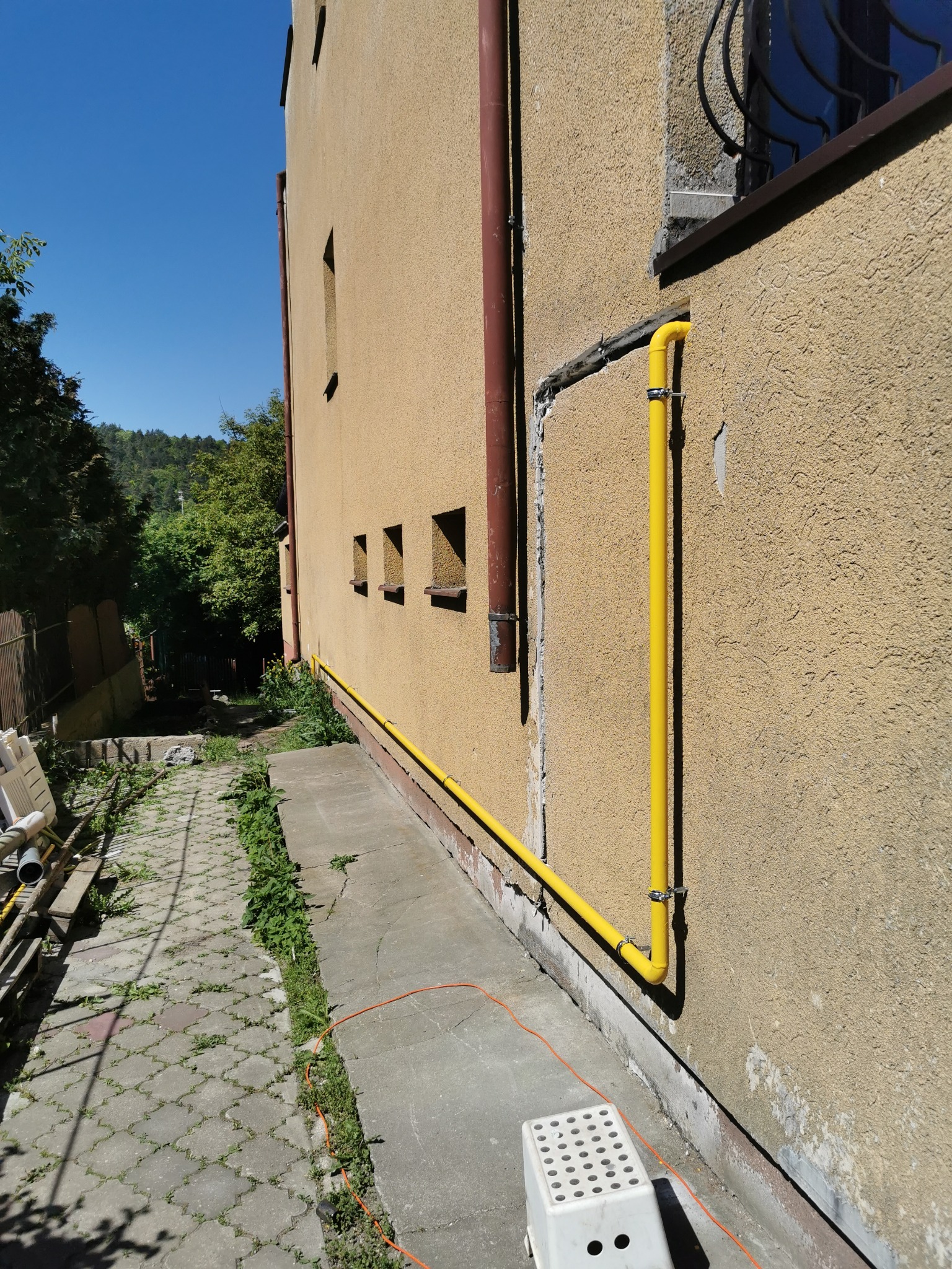 Żółta rura gazowa przymocowana do ściany budynku z fakturą, widoczna z boku, z elementami zieleni w tle i fragmentem chodnika z kostki brukowej.