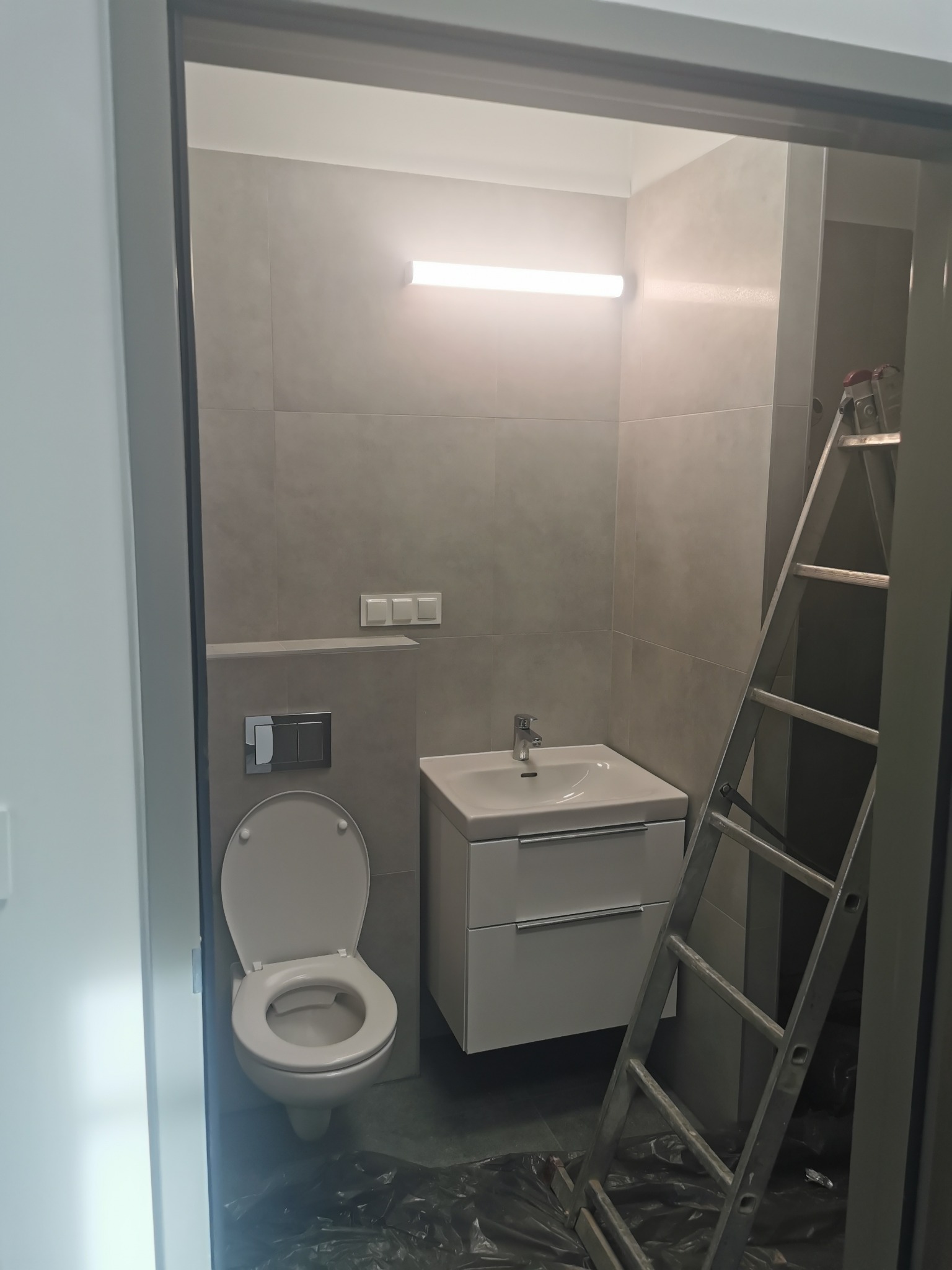 Wykończona łazienka w szarościach z podwieszaną szafką z umywalką, toaletą i drabina oparta o ścianę. Widok zza futryny drzwi.