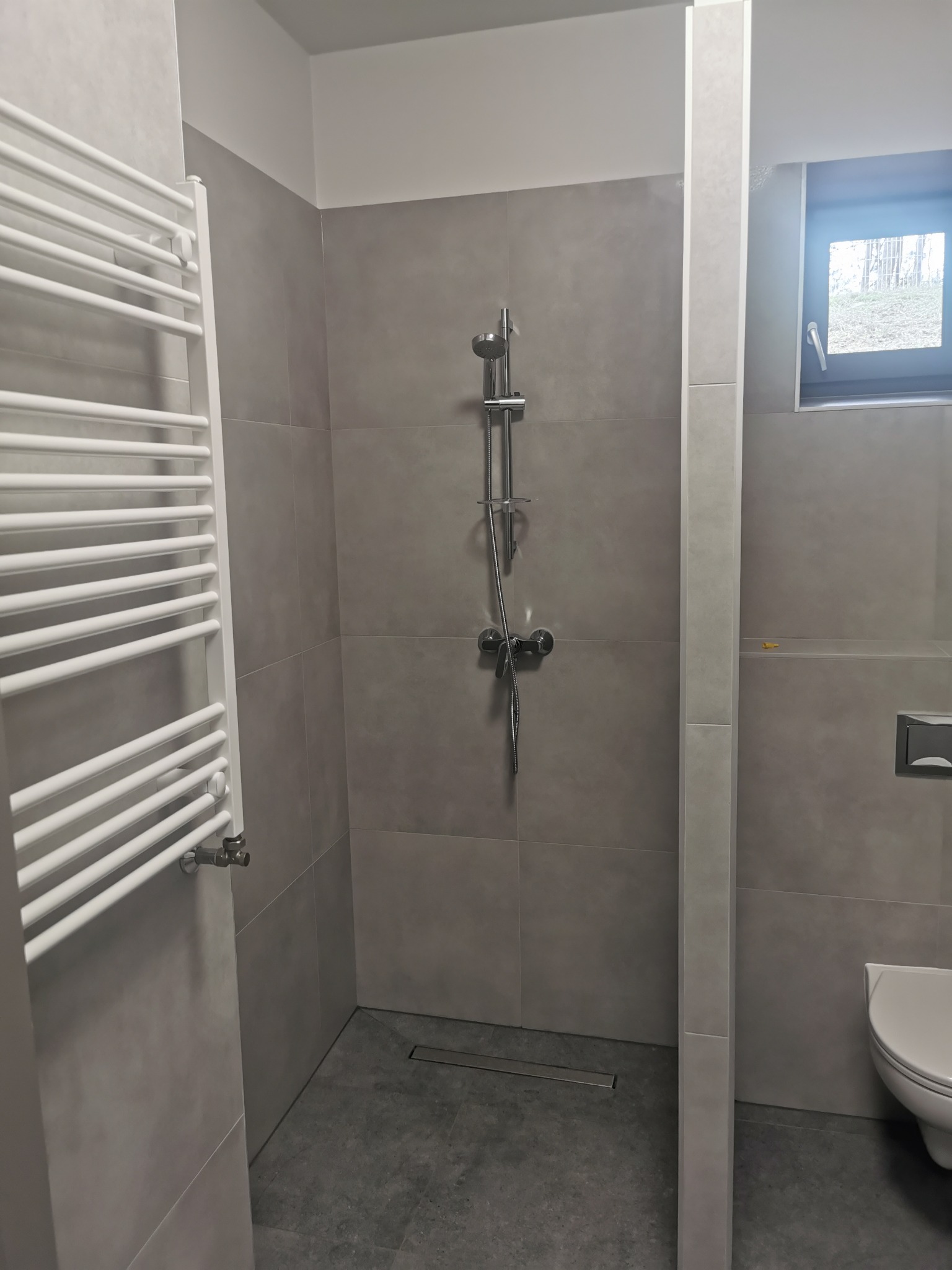 Nowoczesna łazienka z szarymi płytkami, grzejnikiem drabinkowym i kabiną prysznicową z odpływem liniowym. Widoczne okno i fragment toalety.