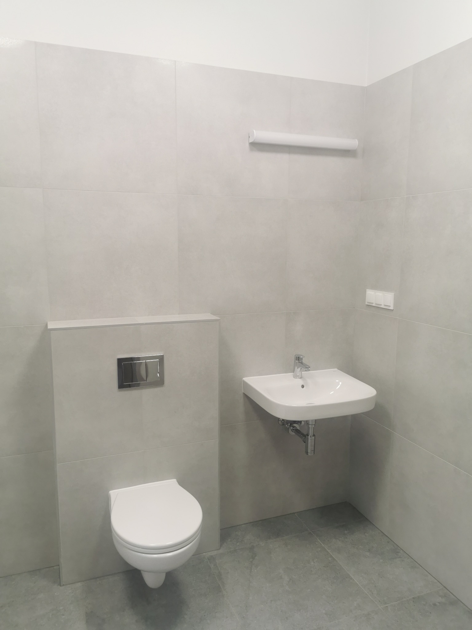 Nowoczesna łazienka w szarościach: podwieszana toaleta z chromowanym przyciskiem, umywalka nablatowa z baterią, szare płytki na ścianach i podłodze.