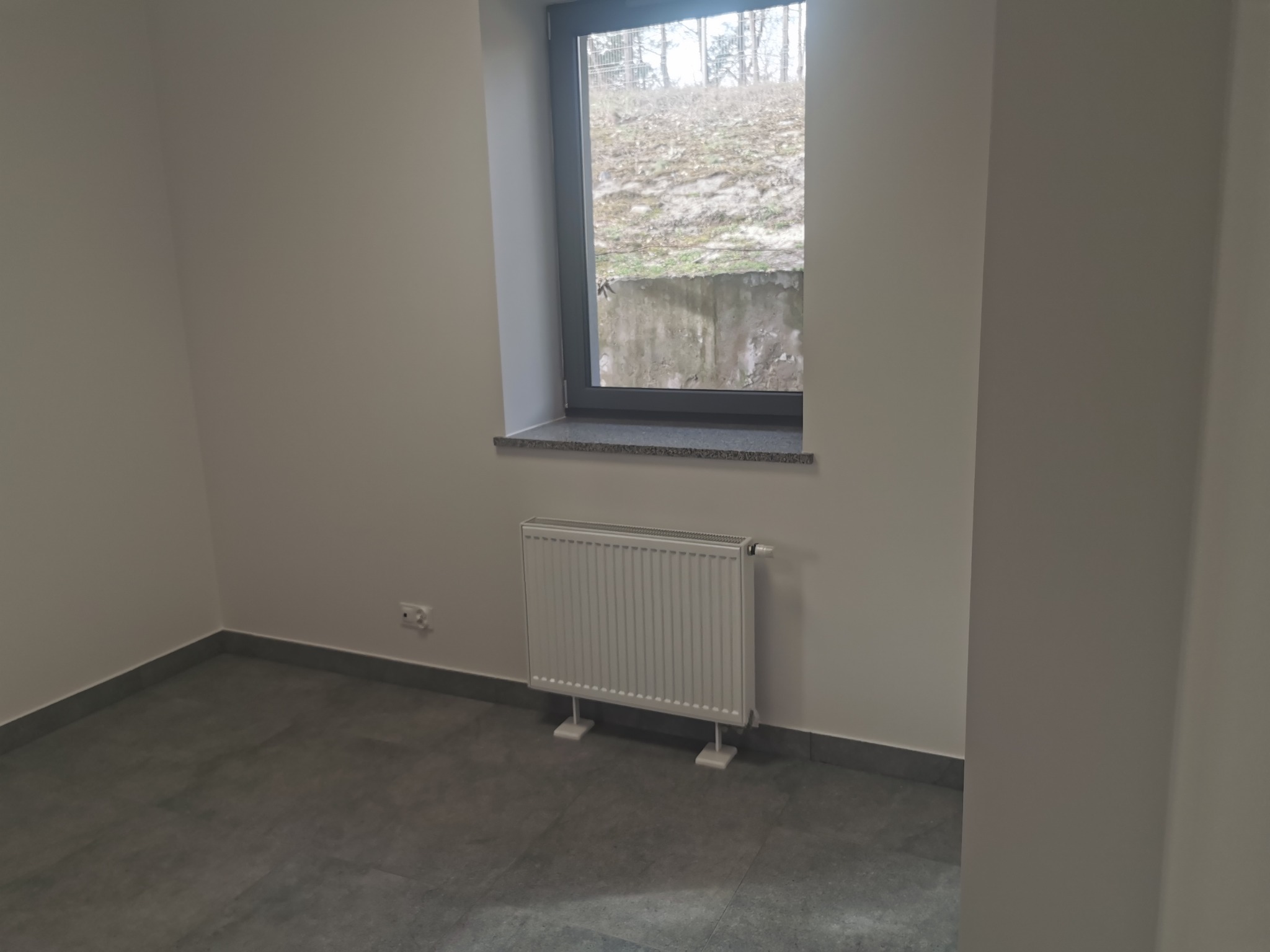 Biały grzejnik panelowy zamontowany pod oknem w jasnym pokoju z szarymi płytkami podłogowymi i widokiem na skarpę. Nowoczesny design, minimalistyczne wnętrze.