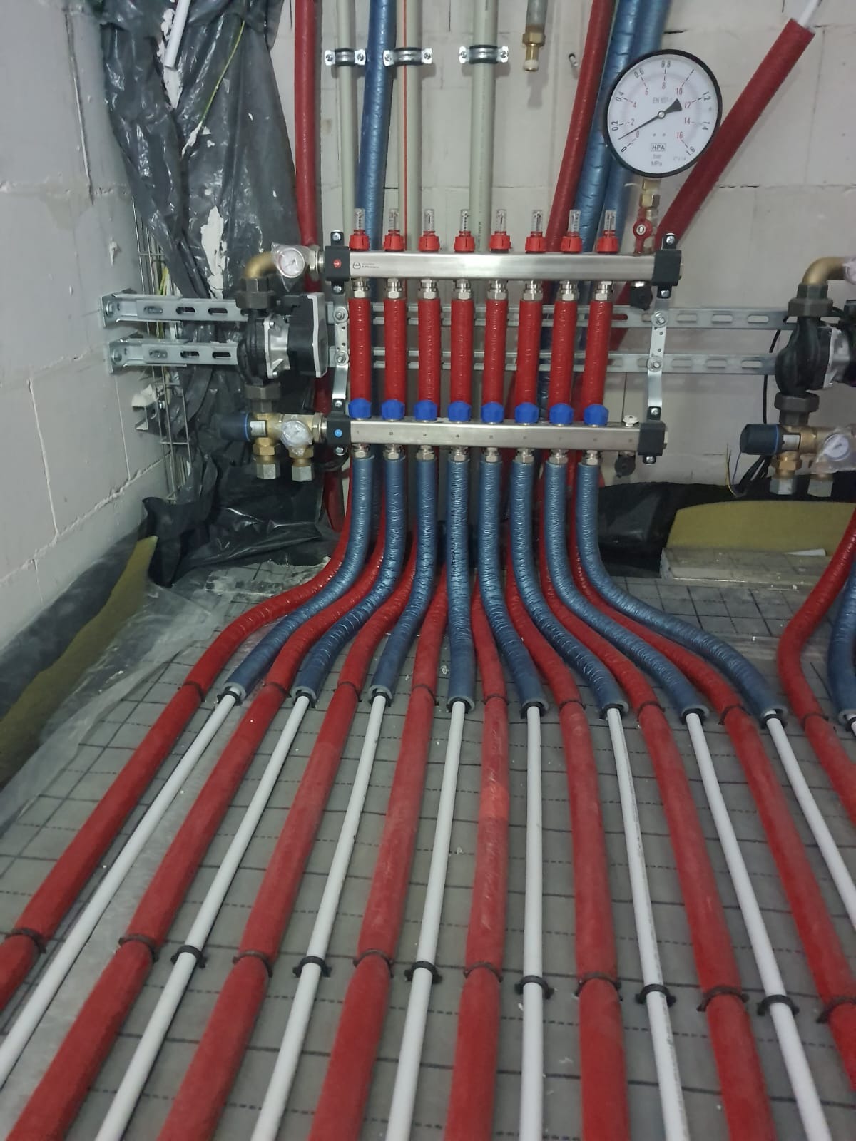 Rozdzielacz ogrzewania podłogowego z rurami w izolacji czerwonej i niebieskiej, zamontowany na ścianie z szarych bloczków, widok z góry na ułożone rury.