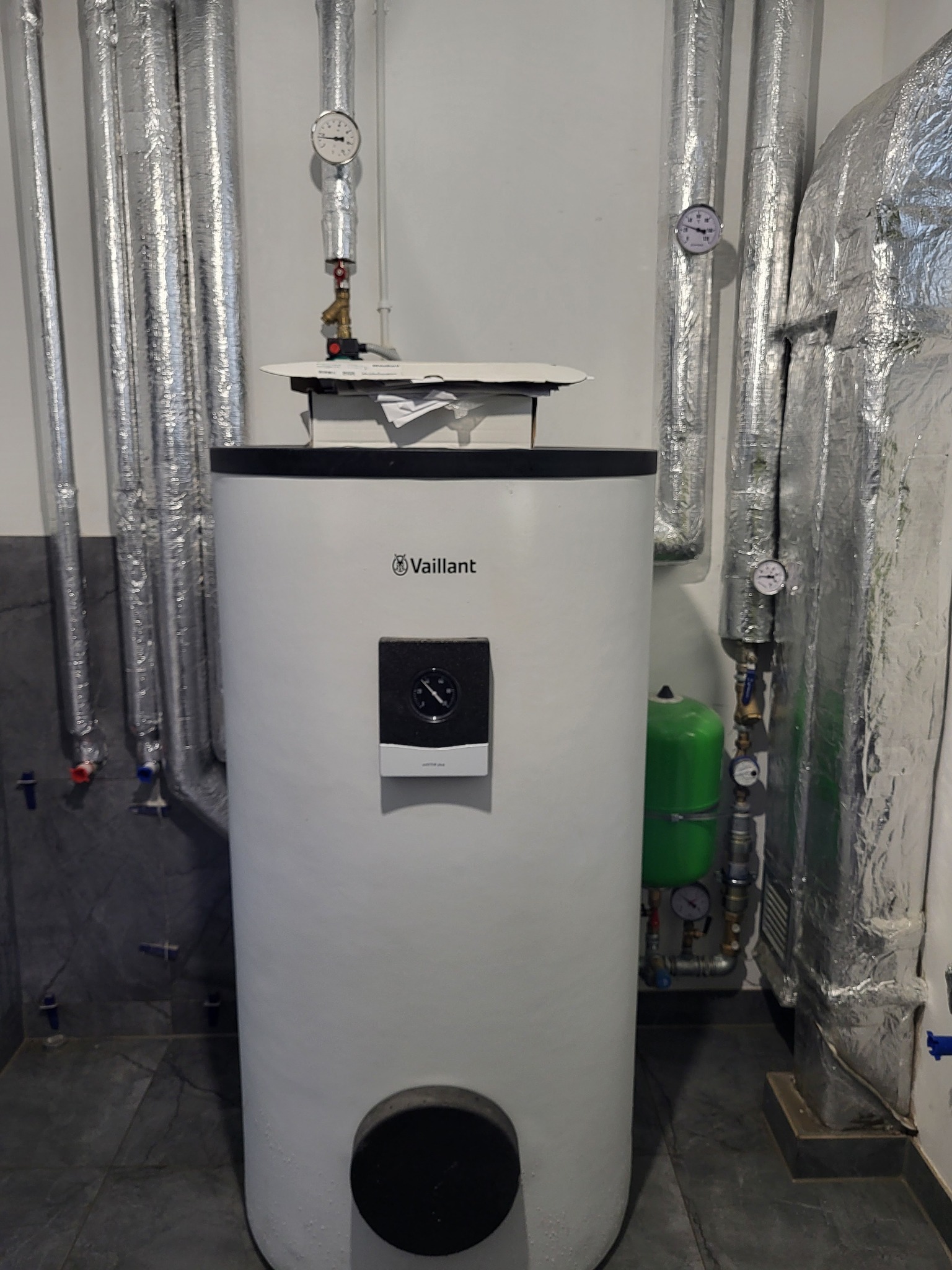 Biały, cylindryczny piec Vaillant z czarnym panelem, otoczony srebrnymi, zaizolowanymi rurami i zielonym zbiornikiem wyrównawczym w kotłowni.