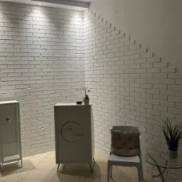 Biała ściana z cegieł w recepcji Ella Studio, z dekoracyjnym ułożeniem. Minimalistyczny wystrój, szklany stolik z rośliną i biała szafka. Jasne wnętrze.