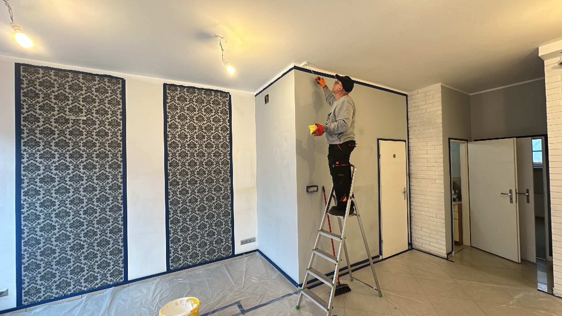 Malarz na drabinie z pędzlem, maluje ścianę przy suficie, zabezpieczoną taśmą. W tle ściana z wzorzystą tapetą, podłoga zabezpieczona folią malarską.