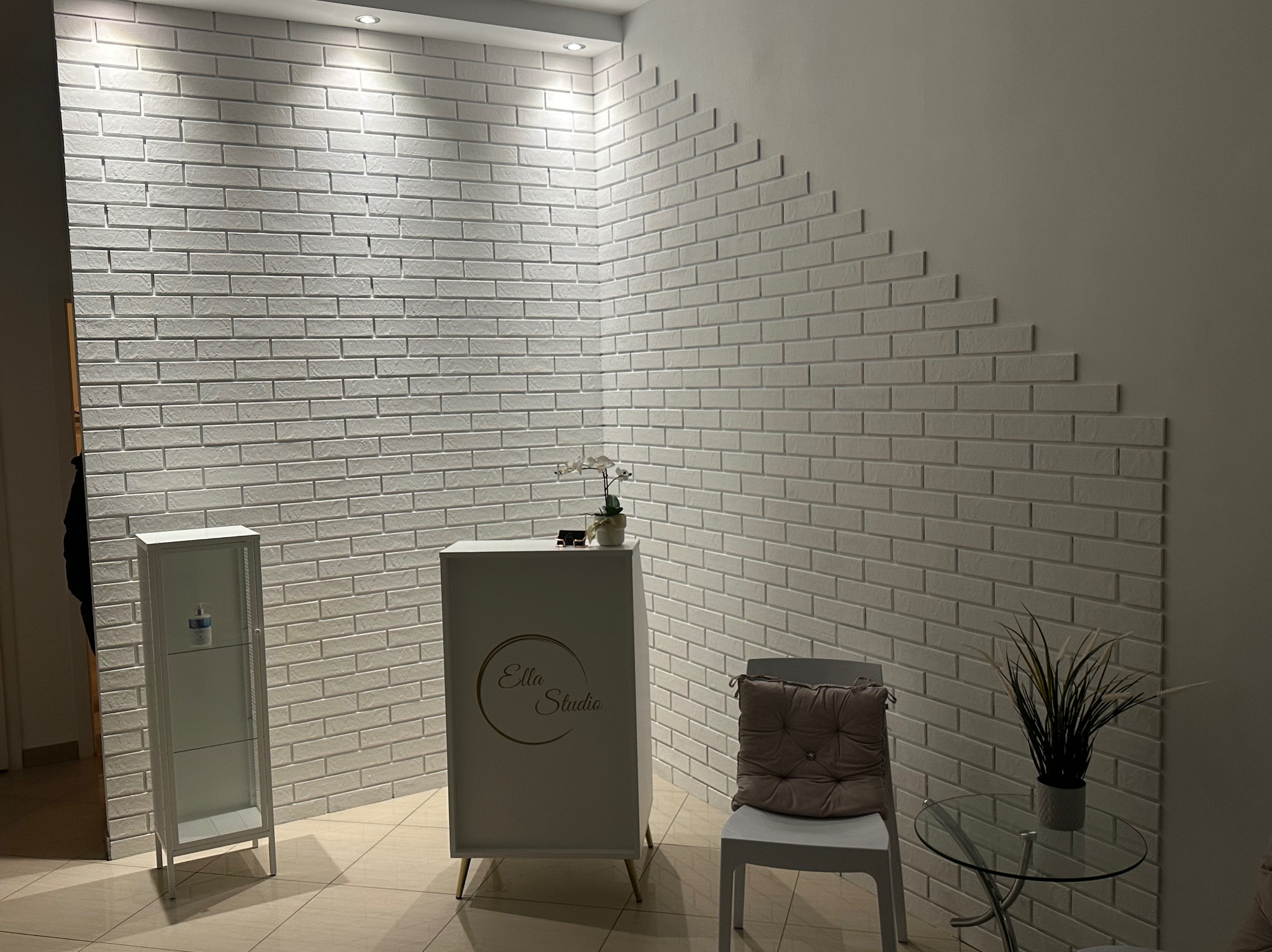 Biała ściana z cegieł w recepcji Ella Studio, z dekoracyjnym ułożeniem. Minimalistyczny wystrój, szklany stolik z rośliną i biała szafka. Jasne wnętrze.