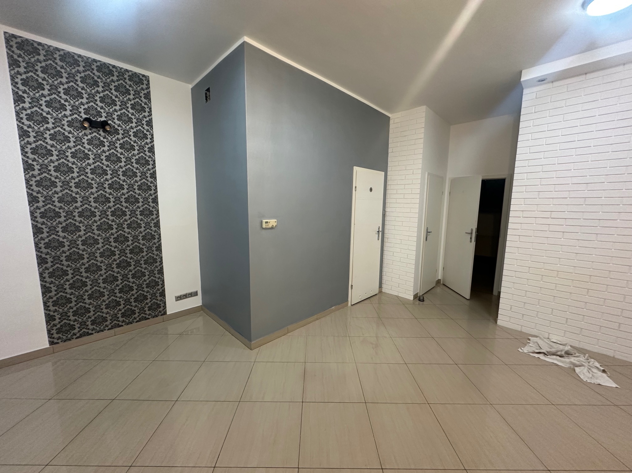 Wnętrze po odświeżeniu: ściana z wzorzystą tapetą, szara ściana z drzwiami i biała ściana imitująca cegłę. Jasne płytki podłogowe. Widok z perspektywy podłogi.
