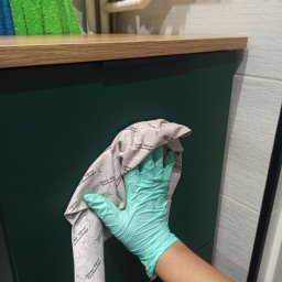 Magic Clean Olena Dudik - Osoba w Lublinie czyści zieloną szafkę ściereczką z mikrofibry w łazience. Widoczna drewniana półka i białe płytki na ścianie.