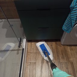 Magic Clean Olena Dudik - Osoba w rękawicach czyści podłogę mopem w łazience z kabiną prysznicową, obok szafka i pralka z ręcznikiem. Widok z góry na proces sprzątania.