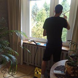 Magic Clean Olena Dudik - Mężczyzna czyści okno w mieszkaniu, używając ściągaczki i żółtego odkurzacza parowego. Widok zza pleców, okno z widokiem na zieleń, roślina doniczkowa w pomieszczeniu.