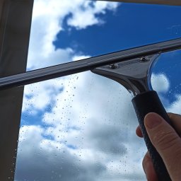 Magic Clean Olena Dudik - Czyszczenie okna z widocznymi kroplami wody i narzędziem do mycia szyb marki Karcher, na tle błękitnego nieba z chmurami.