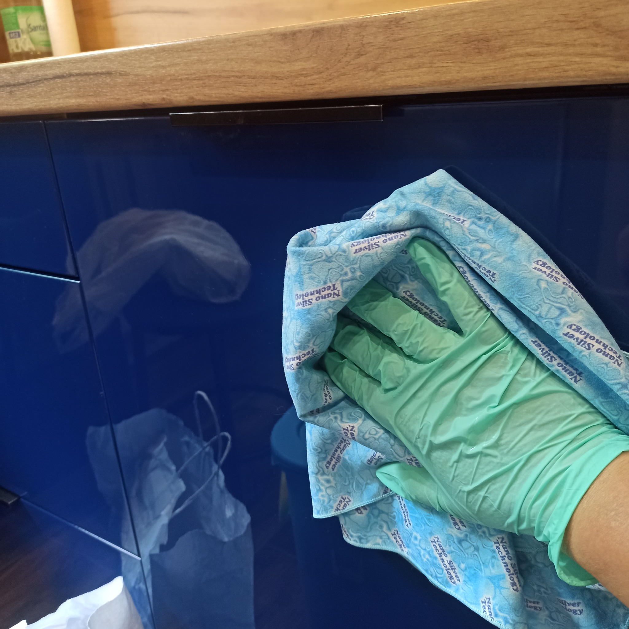 Czyszczenie niebieskich, lakierowanych frontów szafek kuchennych ściereczką z mikrofibry w rękawicy ochronnej. Widoczne refleksy światła.