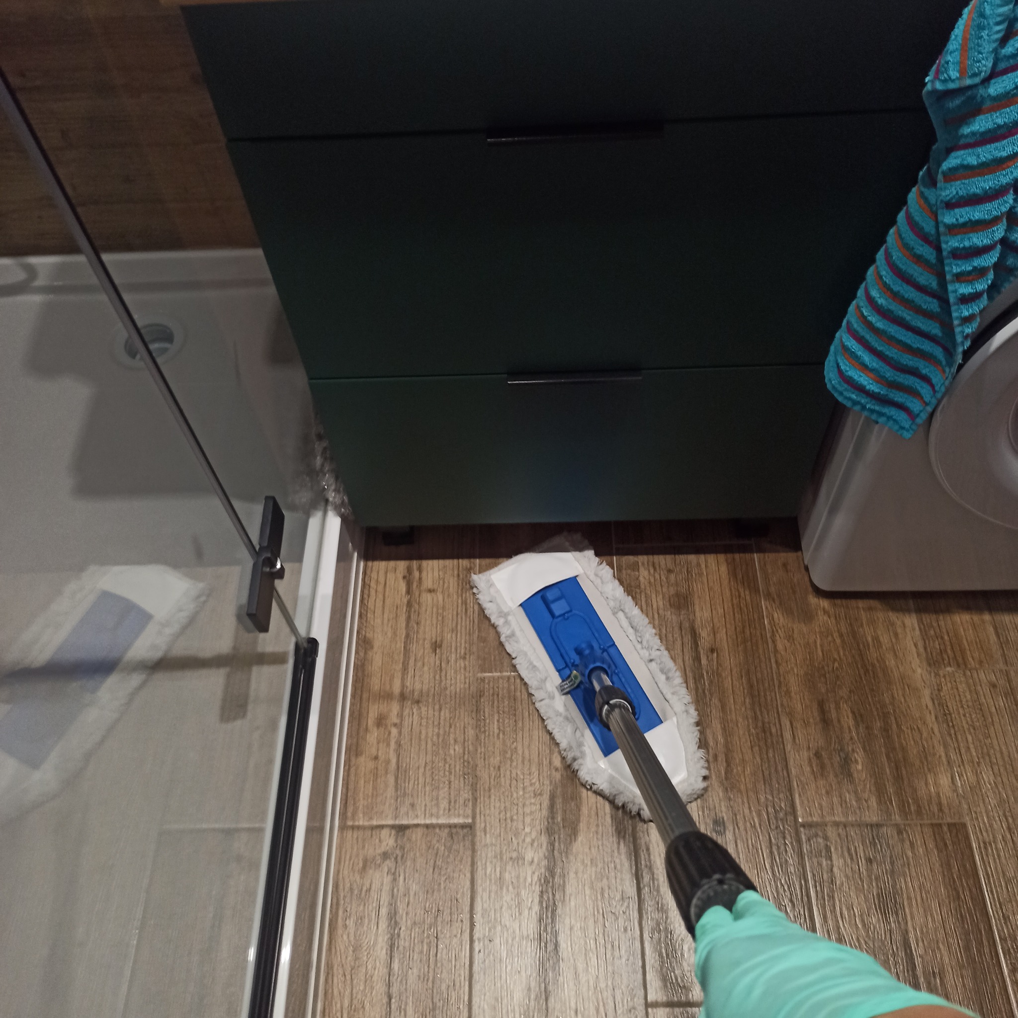 Osoba w rękawicach czyści podłogę mopem w łazience z kabiną prysznicową, obok szafka i pralka z ręcznikiem. Widok z góry na proces sprzątania.