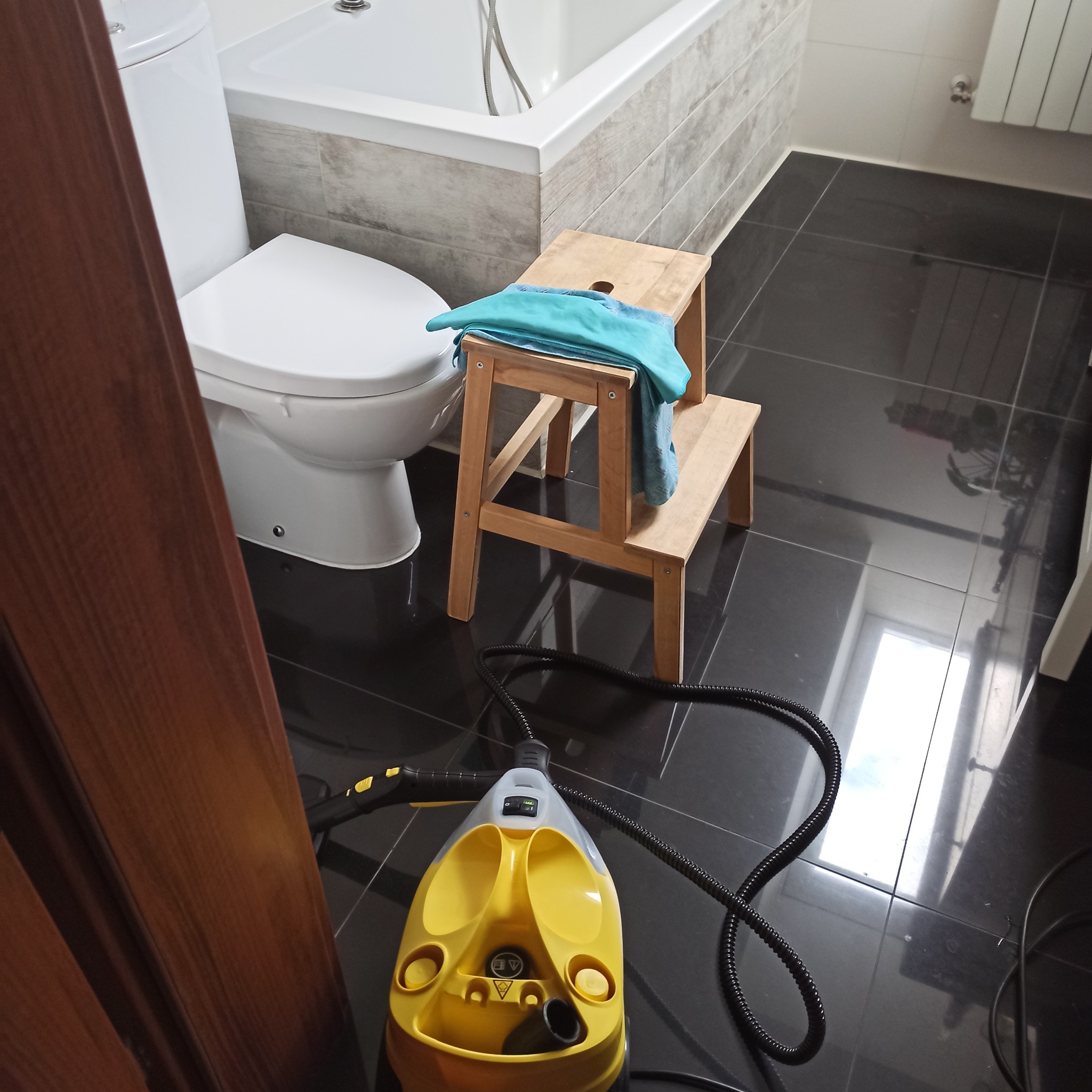 Żółty parowy mop na czarnych płytkach w łazience, obok toaleta i drabinka z ręcznikiem. Widok z góry, jasne światło odbija się od podłogi.