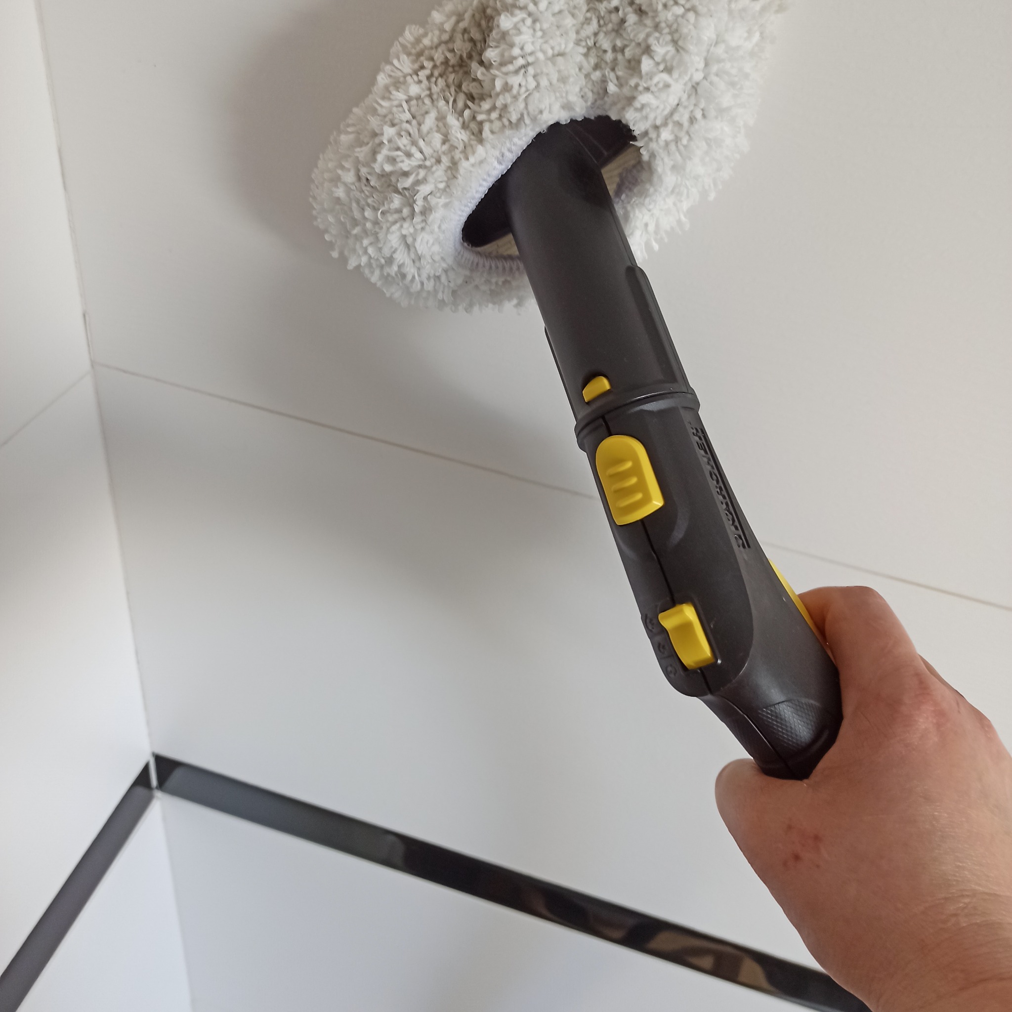 Czyszczenie parowe białej, błyszczącej ściany z czarnymi akcentami przy użyciu parownicy z nakładką z mikrofibry. Widoczna dłoń trzymająca urządzenie.