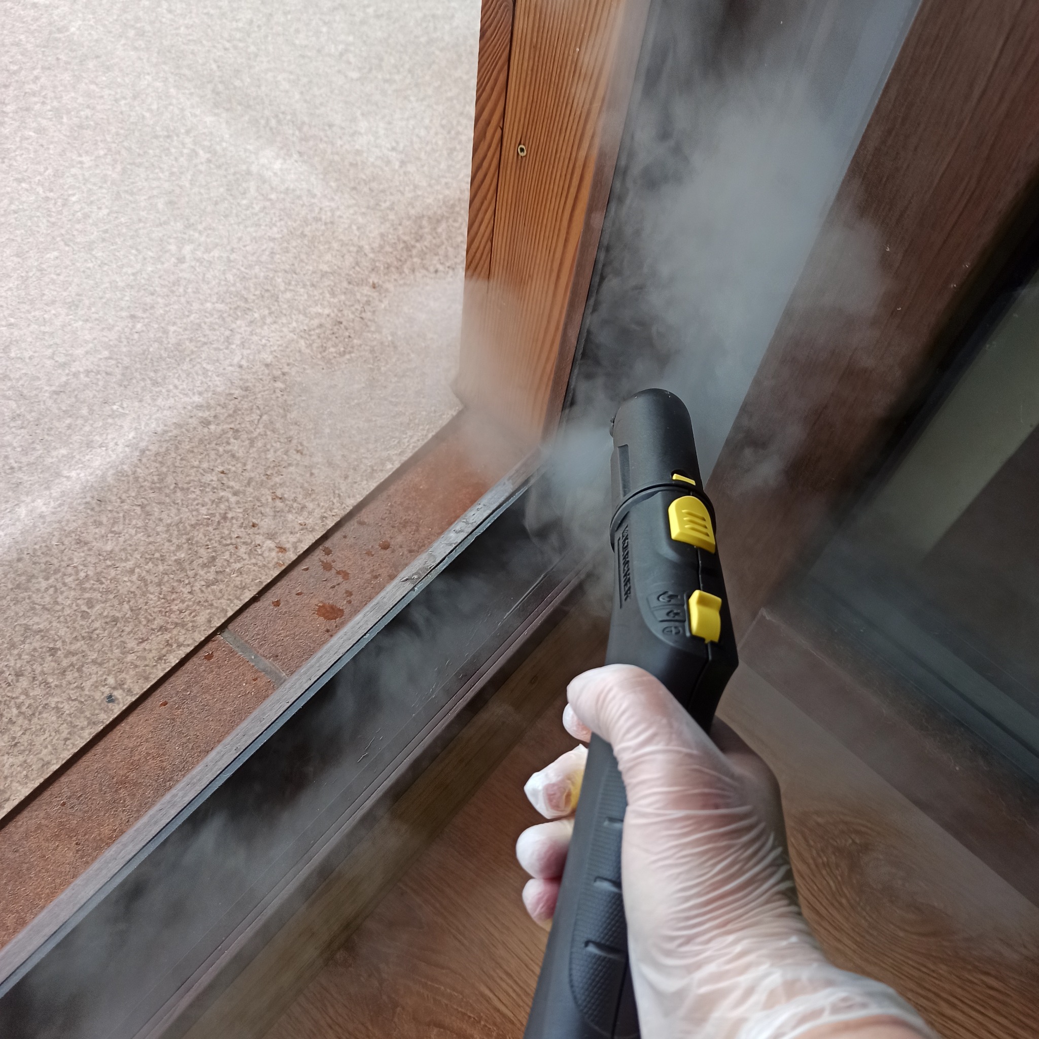 Czyszczenie parowe ramy okiennej z użyciem parownicy Karcher. Dłoń w rękawicy ochronnej trzyma dyszę, z której wydobywa się para. Widoczne drewniane elementy ramy okiennej.
