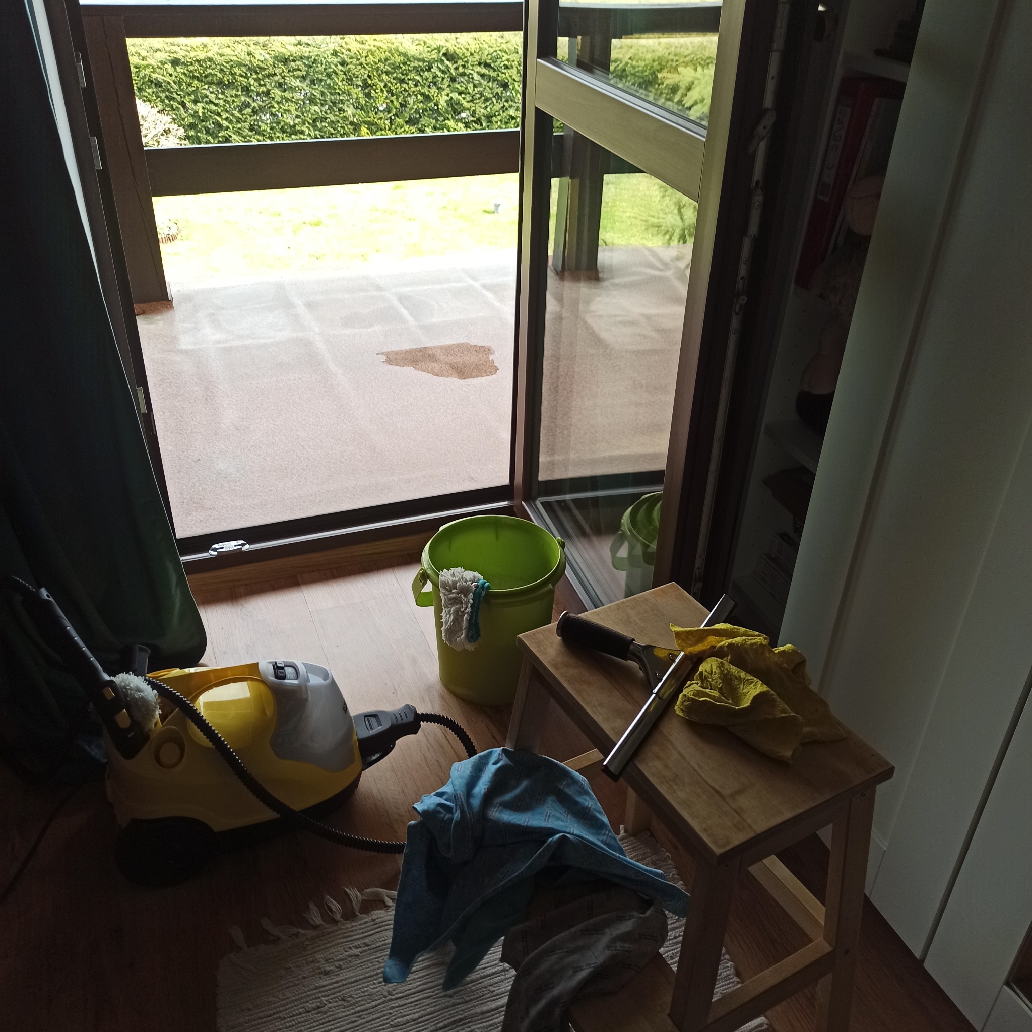 Sprzęt do mycia okien i podłóg: parownica, wiadro z mopem, ściągaczka i ściereczki na tle rozsuwanych drzwi balkonowych z widocznym zabrudzeniem.