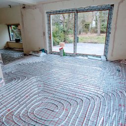 Juliusz Wawrzysz - Białe rury ogrzewania podłogowego ułożone w pętle na czerwonej macie w jasnym, remontowanym pokoju z dużym oknem balkonowym i widokiem na ogród.