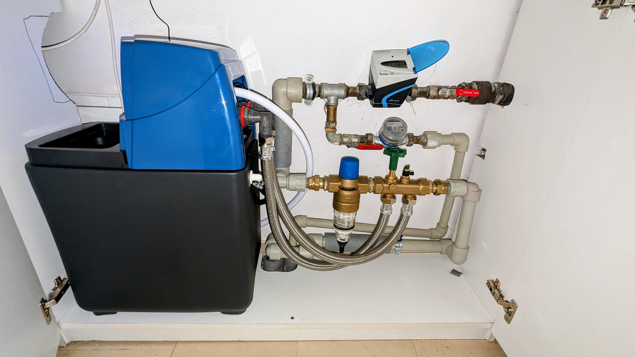 Niebiesko-czarny zmiękczacz wody z podłączeniami hydraulicznymi w szafce. Widoczne rury, zawory, filtr i licznik. Instalacja wykonana w białym wnętrzu szafki.