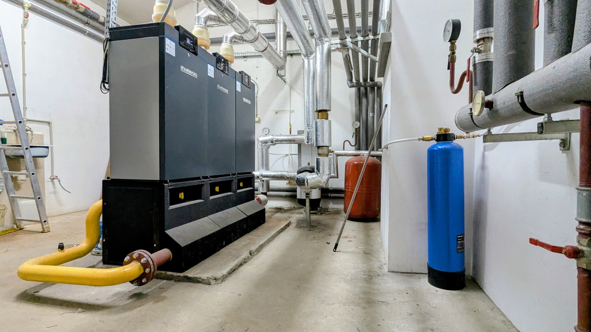Kompleksowa kotłownia z trzema kotłami Buderus, rurami, zbiornikiem wyrównawczym i filtrem wody. Żółta rura gazowa. Instalacja w pomieszczeniu technicznym.