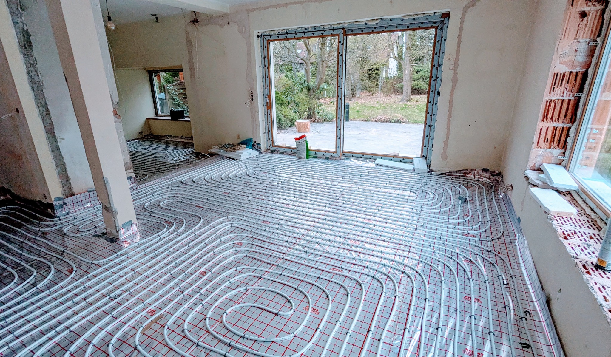 Białe rury ogrzewania podłogowego ułożone w pętle na czerwonej macie w jasnym, remontowanym pokoju z dużym oknem balkonowym i widokiem na ogród.