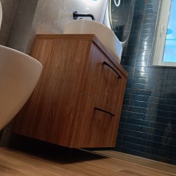 VOKMIS BUD SERHII SIMKOV - Nowoczesna łazienka z drewnianą szafką pod umywalką, okrągłym lustrem z podświetleniem LED i ciemnymi płytkami na ścianie. Widok z niskiego kąta.